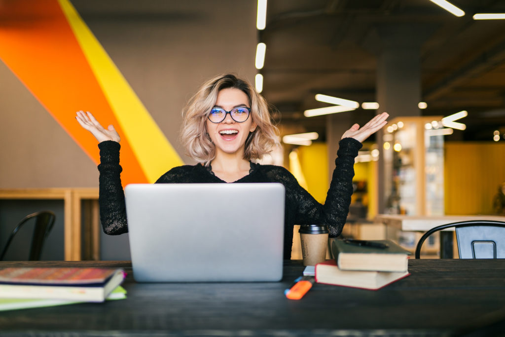 Funny Happy Excited Young Pretty Woman Sitting At Table In Black Shirt Working On Laptop In Co Working Office, Wearing Glasses - Nuvecon